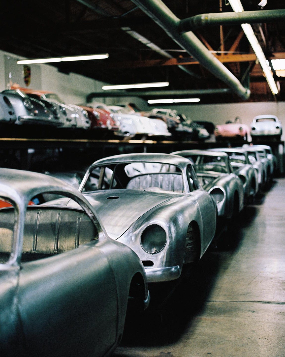 A line of unpainted car bodies sits on a garage floor with more finished cars on a shelf in the background.