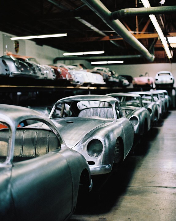 A line of unpainted car bodies sits on a garage floor with more finished cars on a shelf in the background.