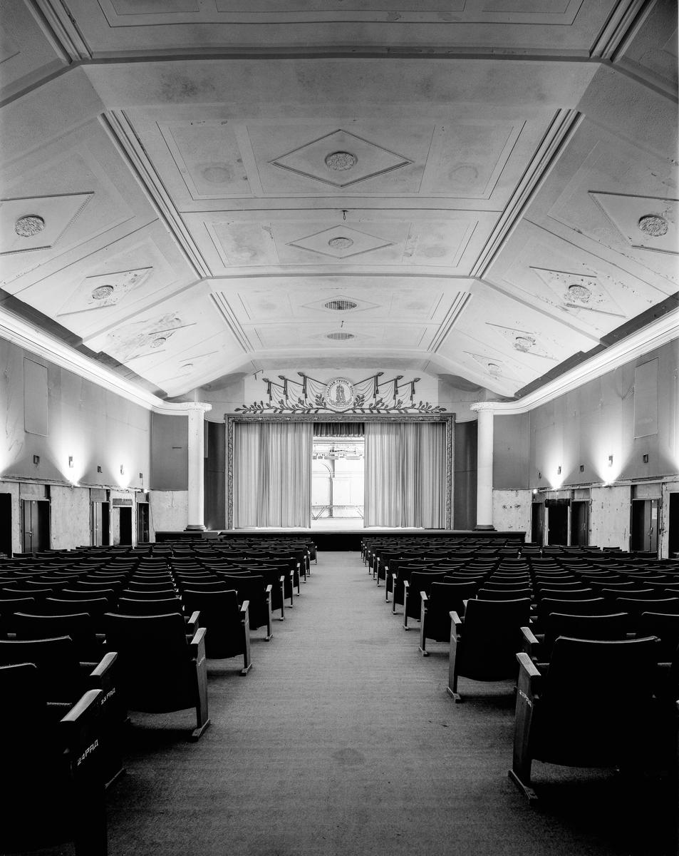 An empty auditorium with rows of seats faces a stage with closed curtains.
