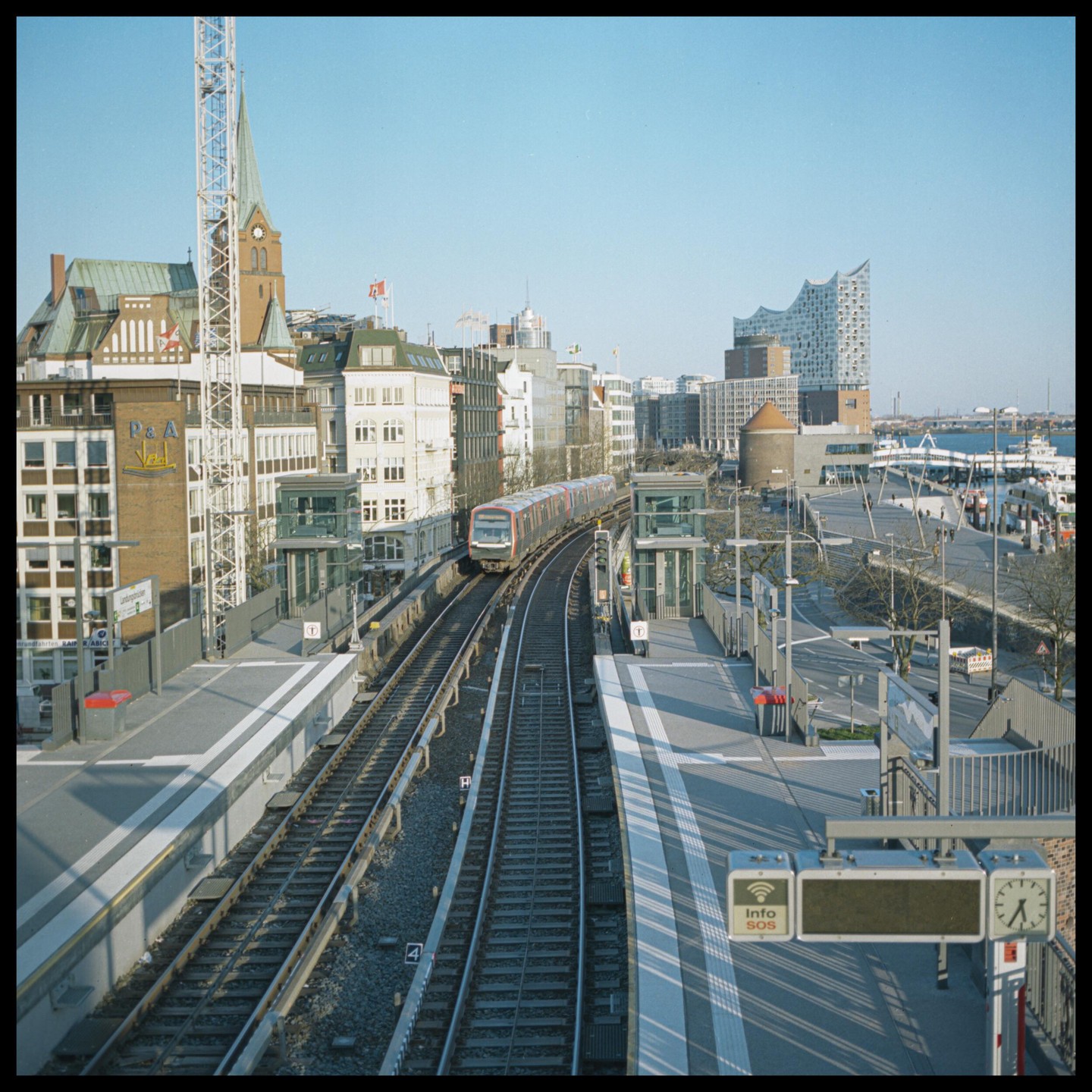 A train approaches a station platform in a city with a harbor and modern buildings.
