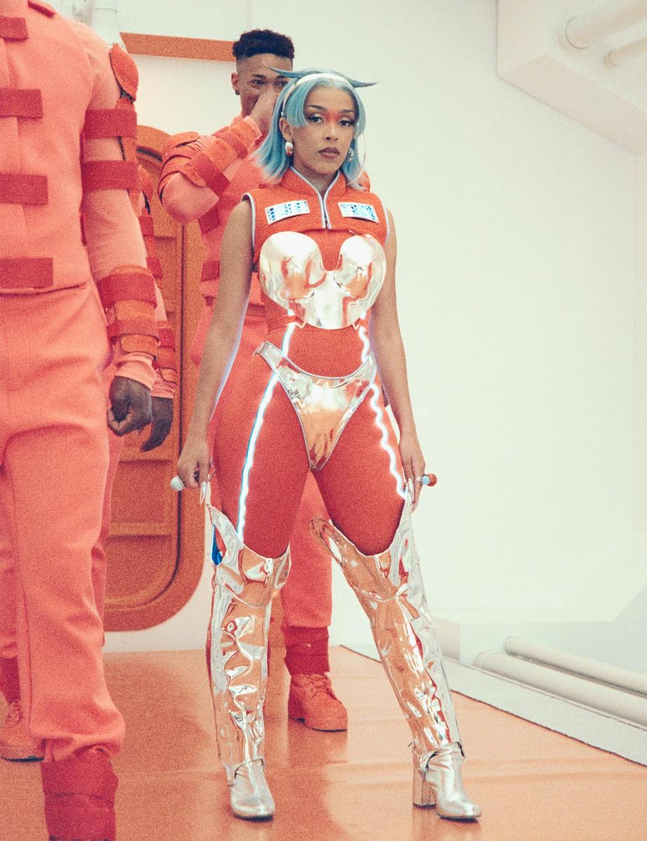 A woman in a futuristic orange and silver outfit stands with two men in orange jumpsuits in a white room.