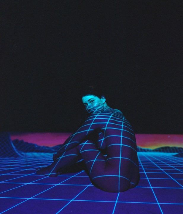 A woman sits on a grid-patterned surface with a grid projected onto her body and a purple and blue landscape in the background.