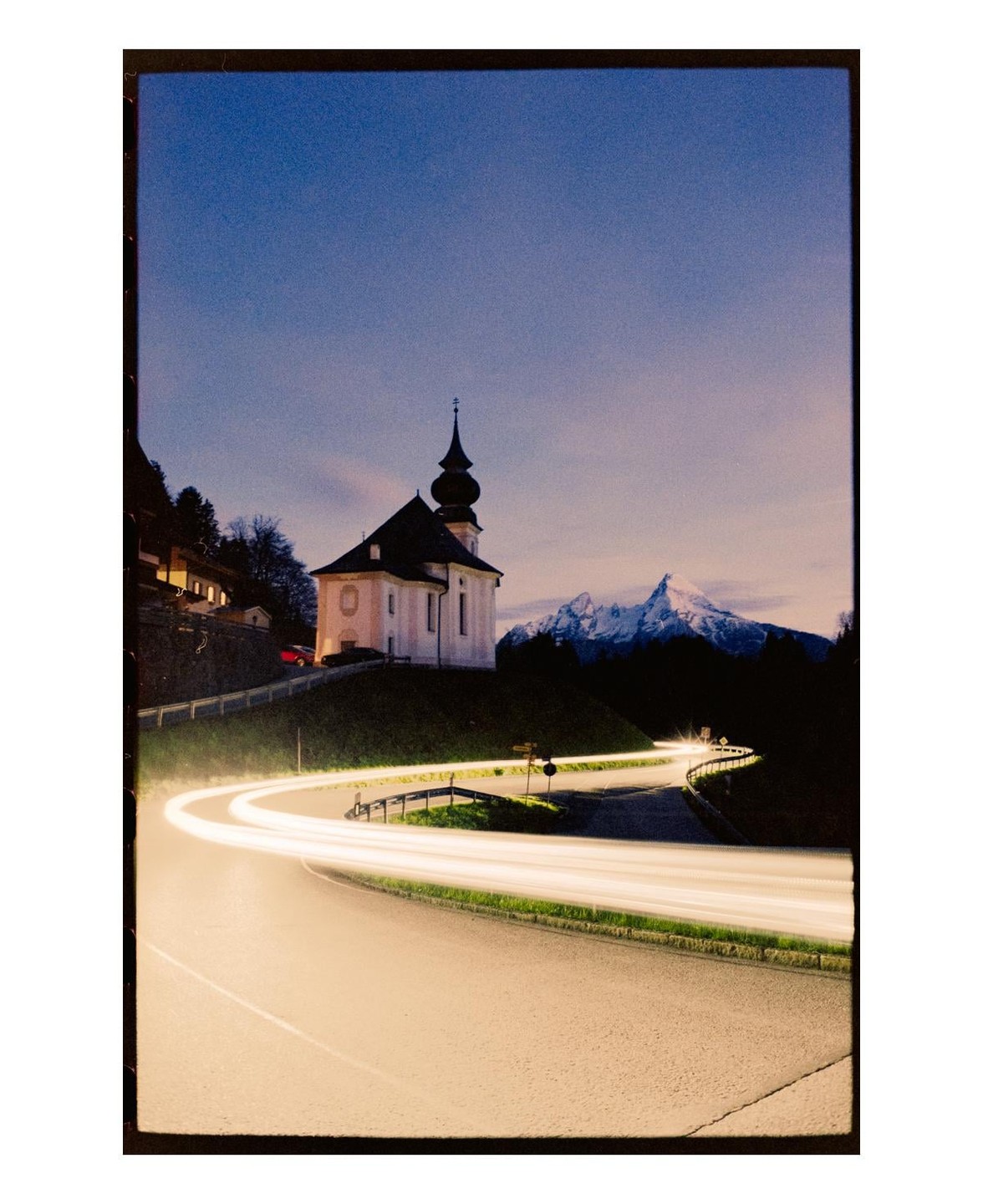 A church sits on a hill overlooking a winding road with light trails and a snow-capped mountain in the background at dusk.