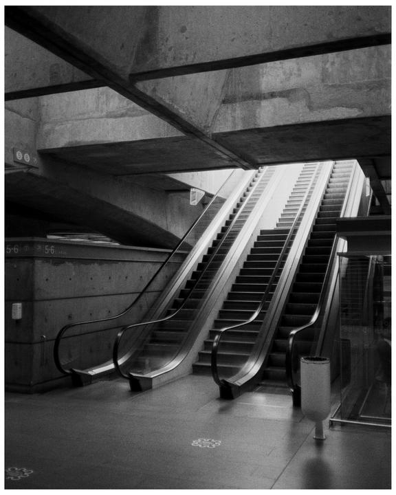 Two escalators and a staircase ascend towards a bright light source in a concrete train station.