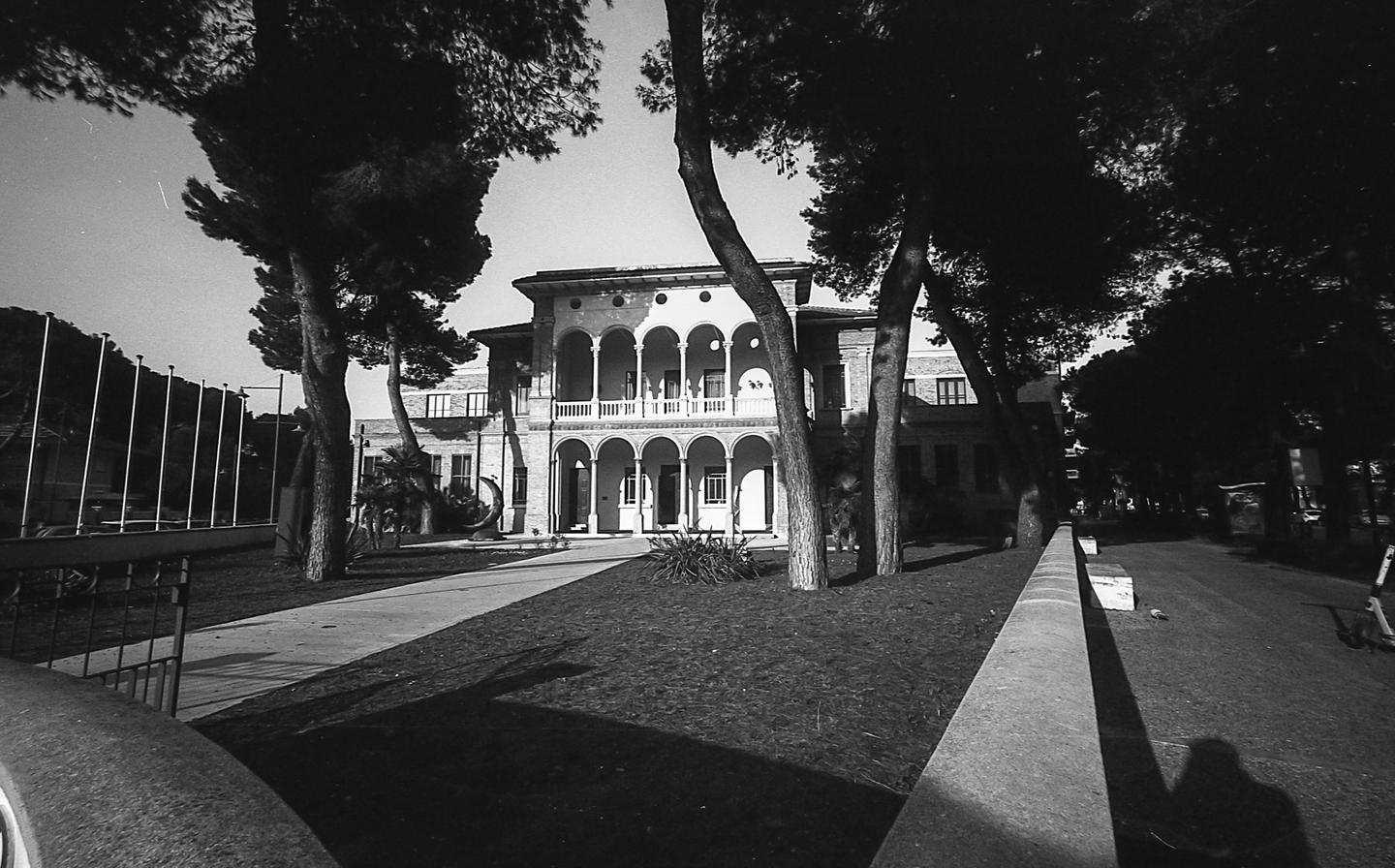 A large building with arches and a balcony is framed by tall trees and a grassy area.