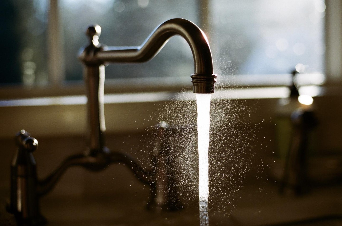 Water streams from a kitchen faucet, illuminated by light from a window.