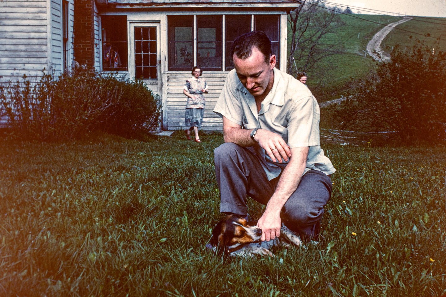A man crouches to pet a dog in the grass while a woman stands on a porch in the background.