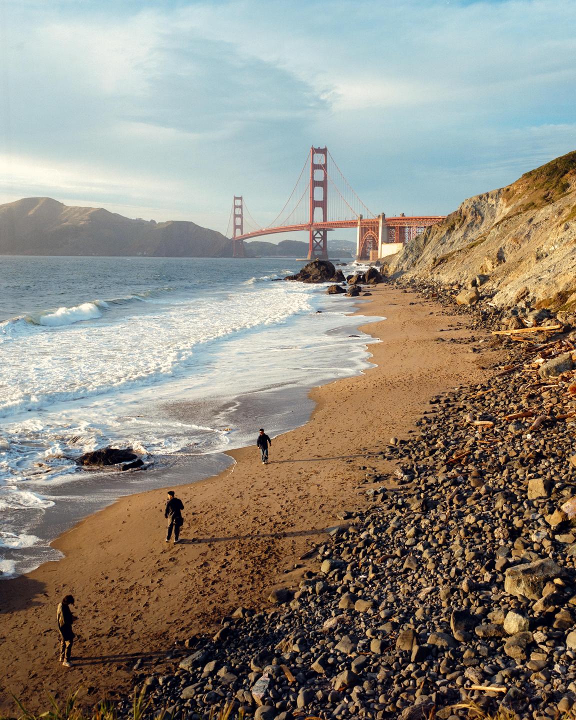 Three people walk along a sandy and rocky beach with the Golden Gate Bridge in the background.