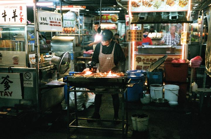A man wearing an apron and a cap is grilling food on skewers over an open flame at a street food stall at night.