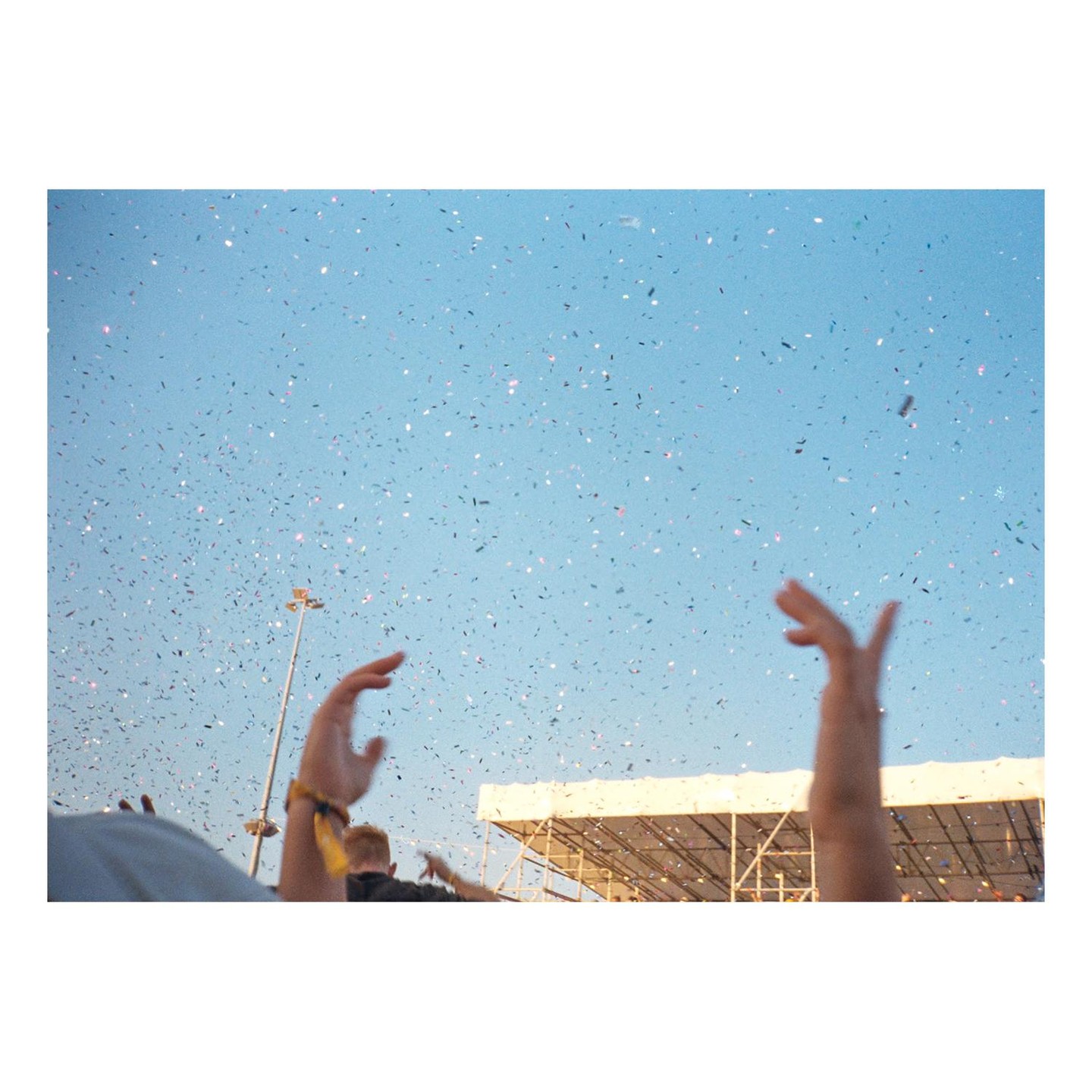 Confetti falls from a clear blue sky over a crowd with raised hands at an outdoor event.