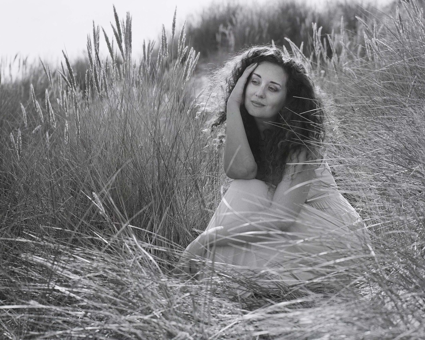 A woman with curly hair sits in tall grass, looking away from the camera.