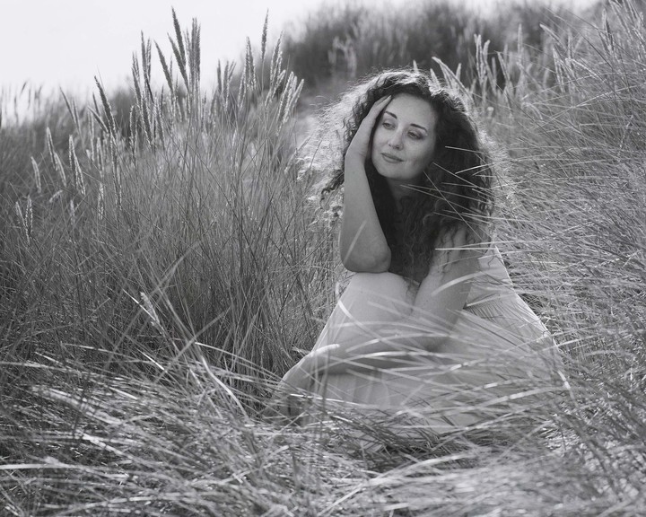 A woman with curly hair sits in tall grass, looking away from the camera.