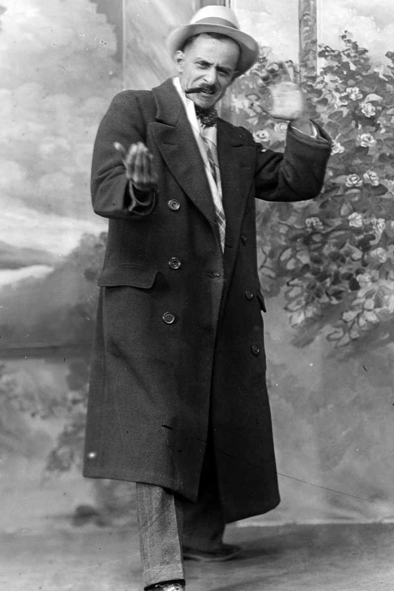 A man in a hat and long coat gestures with his hands while smoking a cigar in front of a painted backdrop.