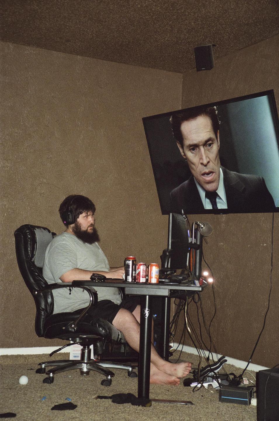 A man with a beard sits at a desk with a computer and several cans, while a large screen on the wall shows a man in a suit.