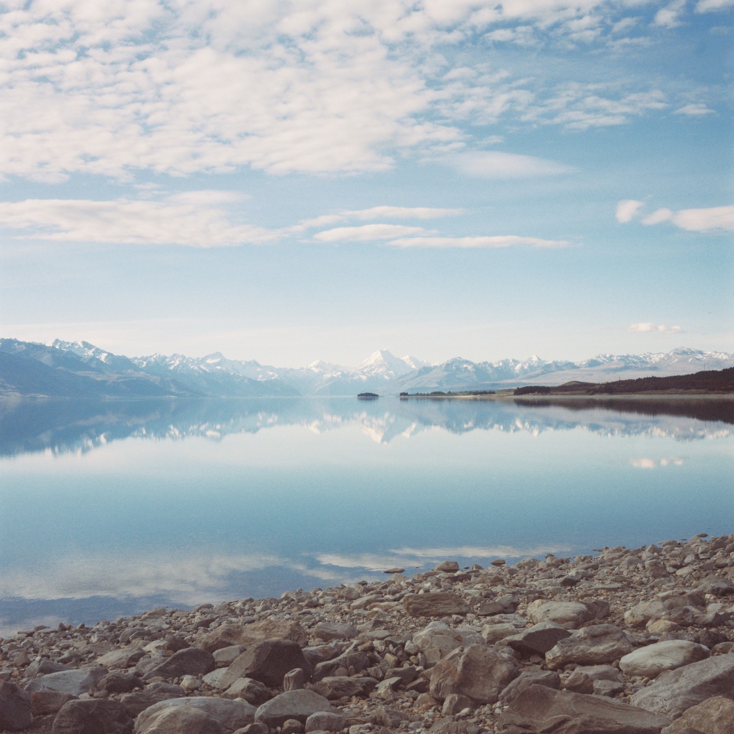 A calm lake reflects snow-capped mountains and a cloudy sky, with a rocky shore in the foreground.