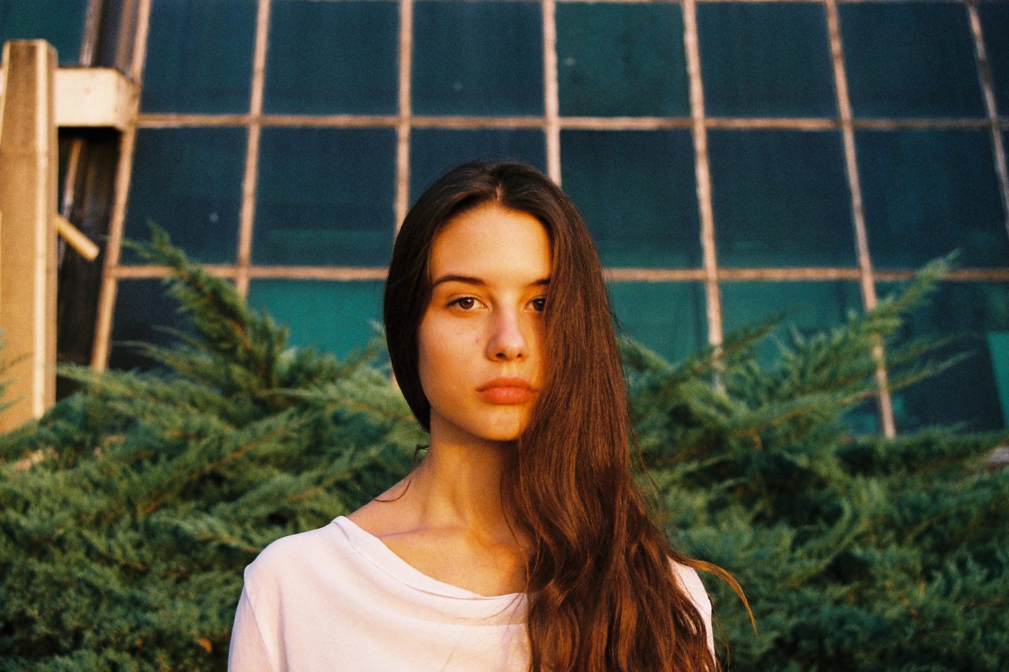 A woman with long brown hair looks directly at the viewer, with green foliage and a building in the background.