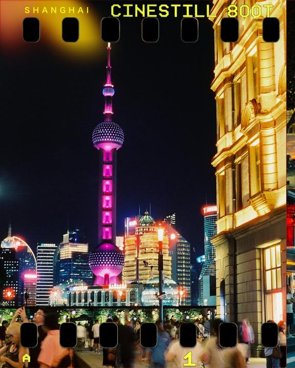 A crowd of people stands on a street at night with a tall, illuminated tower and other buildings in the background.