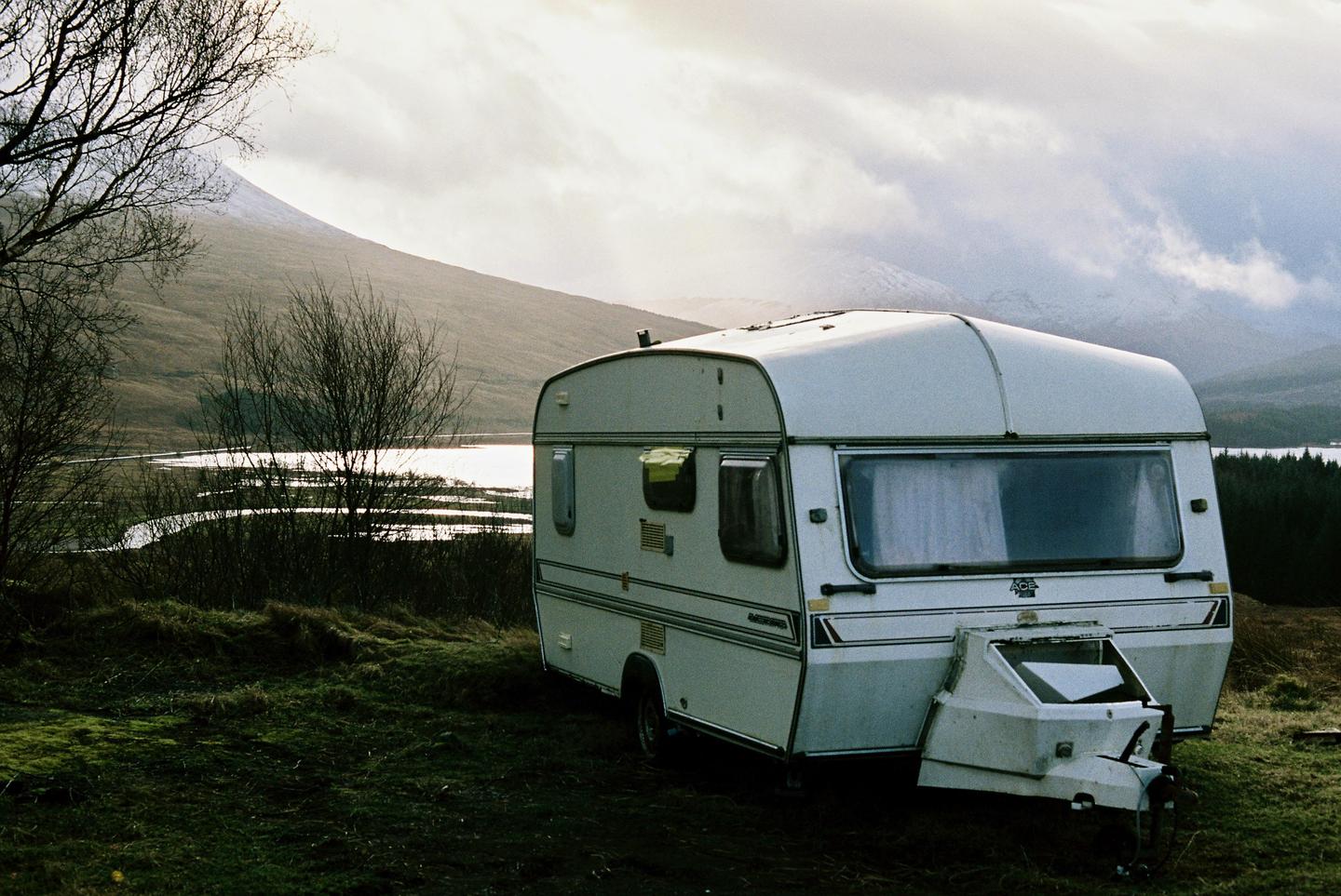A white caravan is parked on a grassy hill overlooking a body of water and mountains under a cloudy sky.