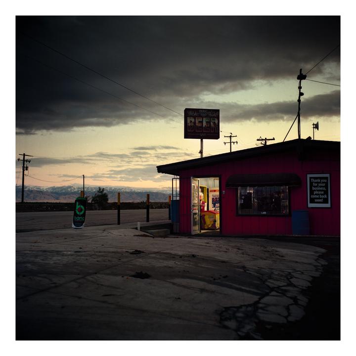 A small red store with an open door and a beer sign stands beside a road with mountains in the distance under a cloudy sky.