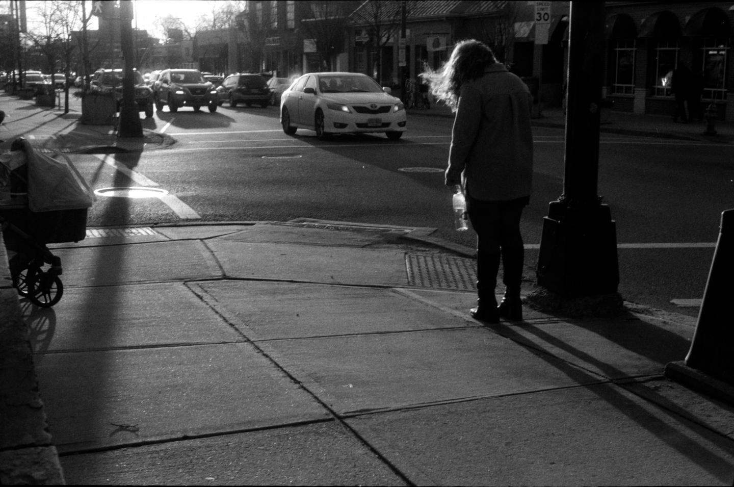 A woman stands on a sidewalk next to a street with cars and a baby carriage.