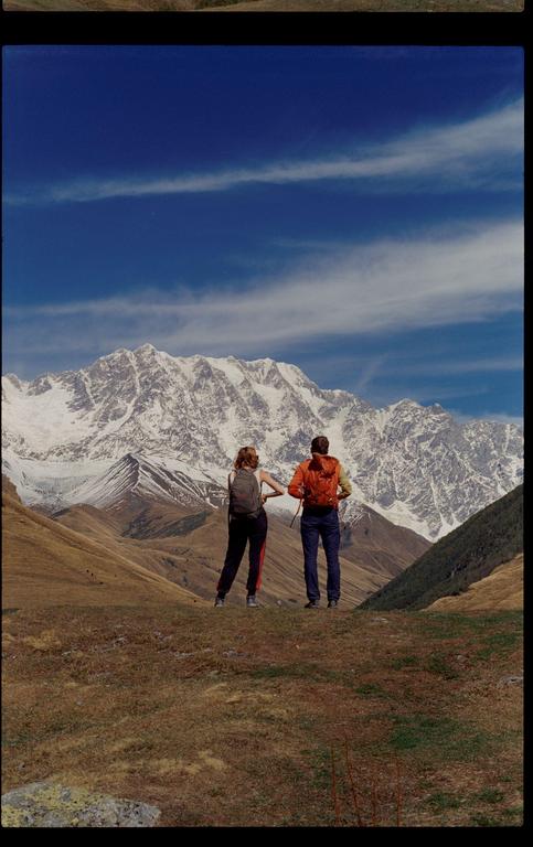 A couple with backpacks stands on a hill overlooking a vast mountain range with snow-capped peaks.