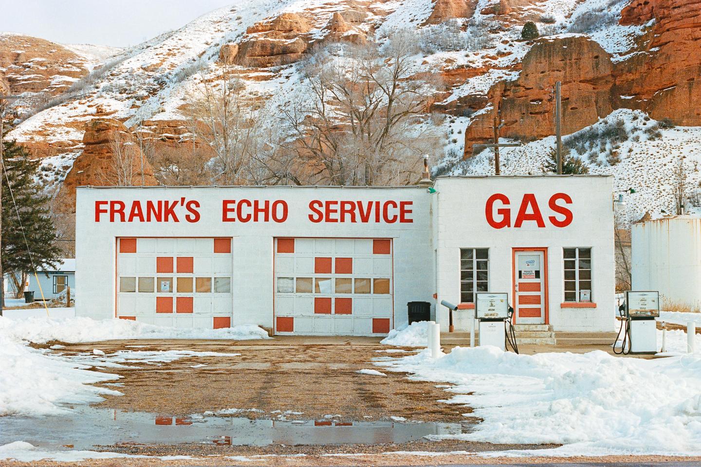 A gas station and service garage with red lettering on a white building stands in a snowy landscape with mountains in the background.