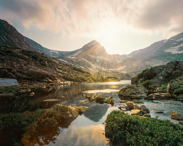 Sunlight streams over a mountain lake surrounded by rocky peaks and green foliage.