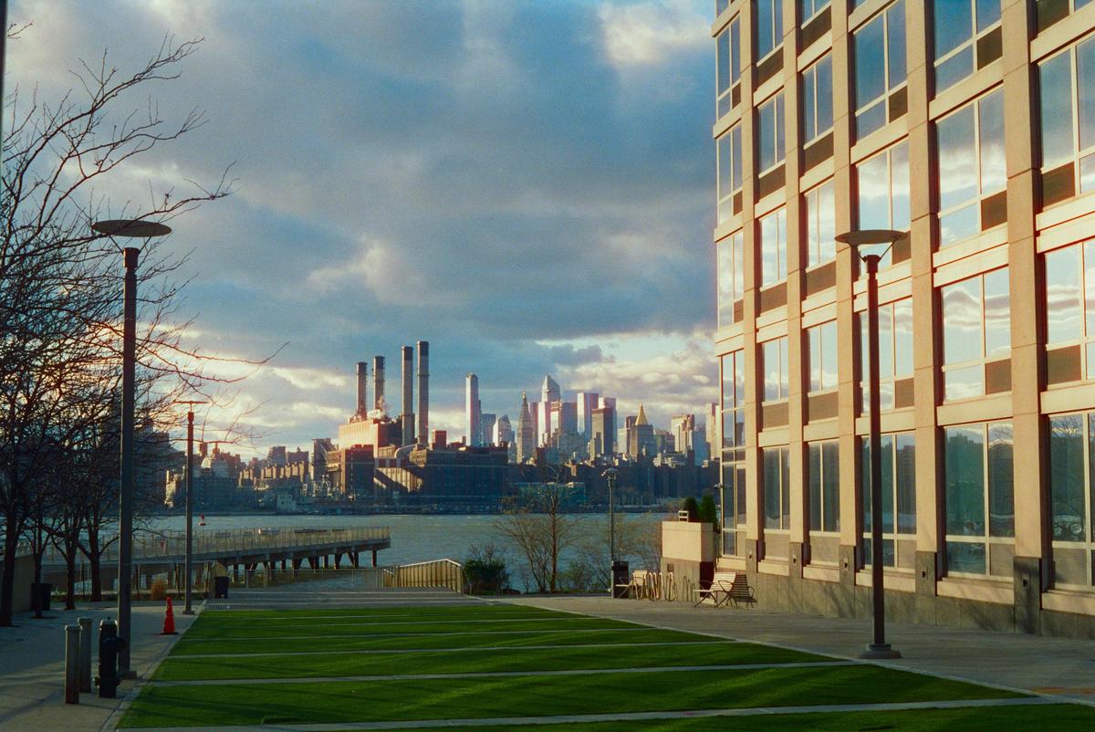 A cityscape with a river in the foreground and a large building on the right, under a cloudy sky.
