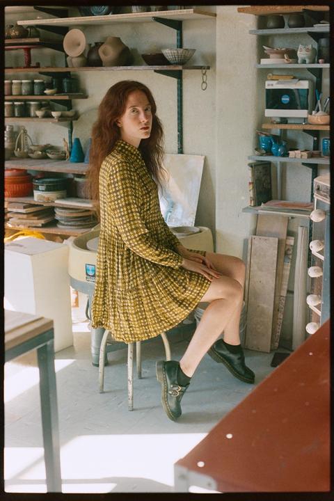 A woman with long red hair sits on a stool in a pottery studio, looking over her shoulder at the viewer.