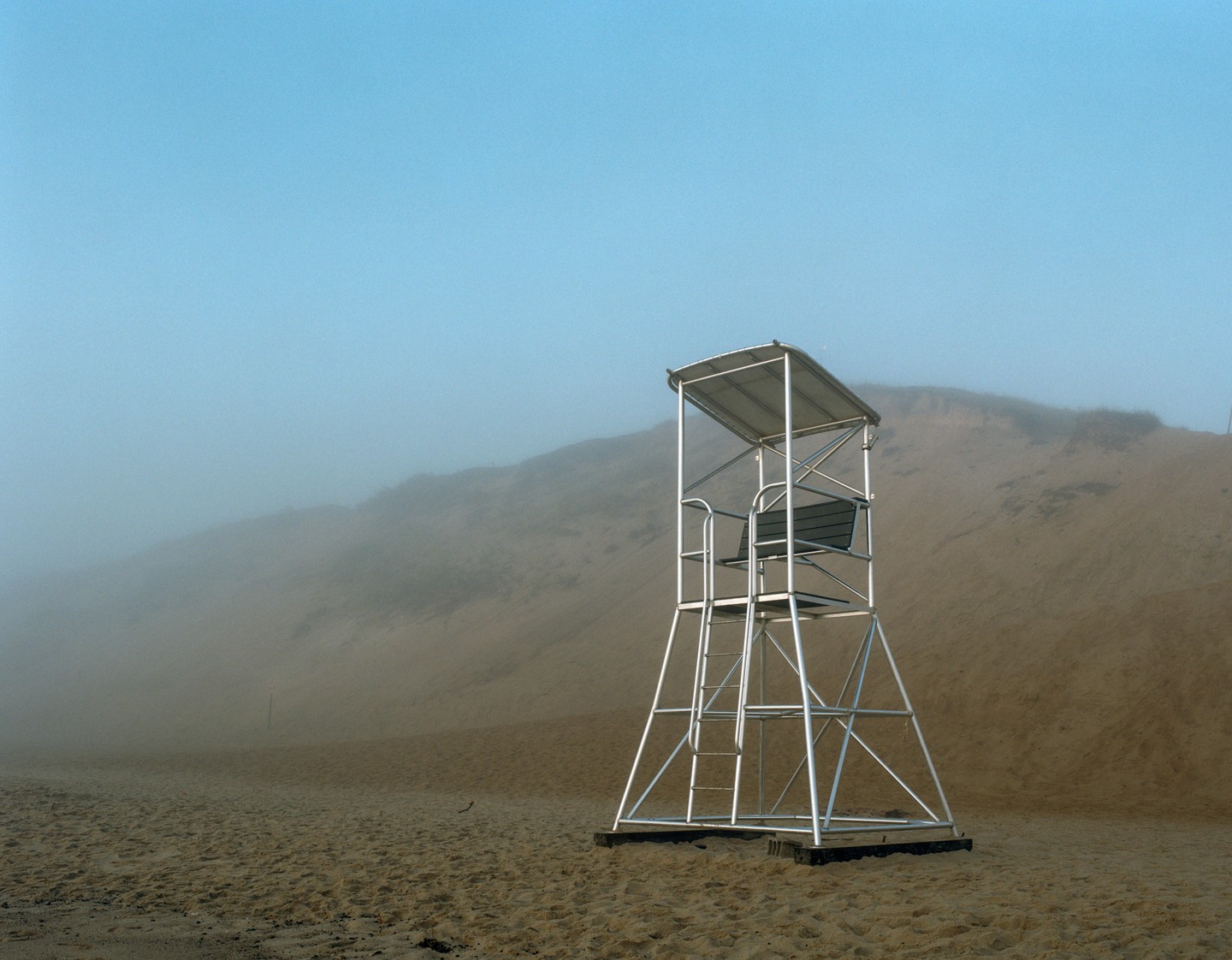 A lifeguard stand sits on a sandy beach with a hazy sky and distant dunes.