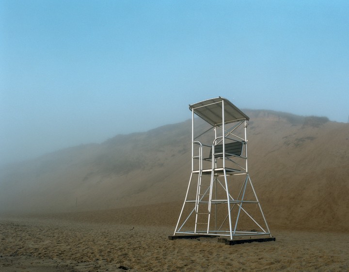 A lifeguard stand sits on a sandy beach with a hazy sky and distant dunes.