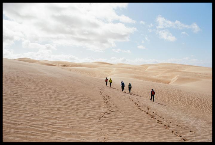 Five people walk in a line across a vast desert landscape under a partly cloudy sky.