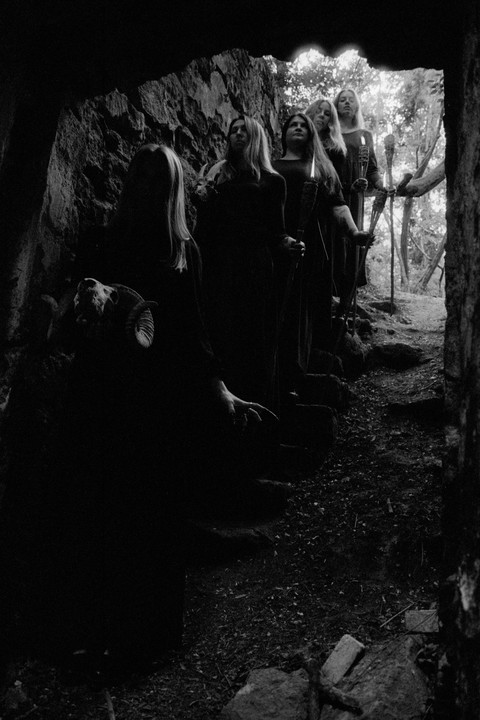 Five women in dark robes stand in a line holding torches, emerging from a dark cave into a forest.