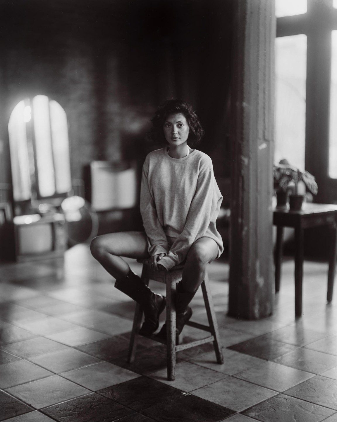 A woman with short, curly hair sits on a wooden stool in a room with tiled floors, wearing a sweatshirt and boots.