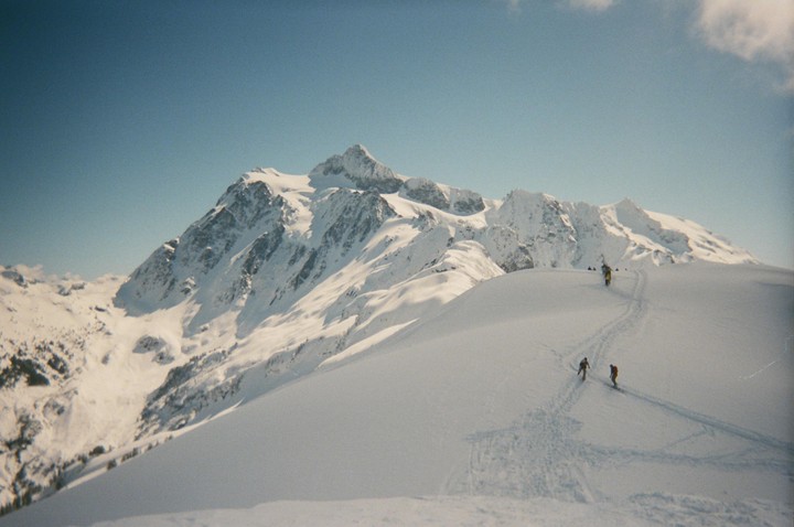 Two skiers descend a snowy mountain with a large snow-covered peak in the background.