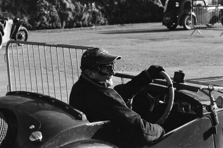A man wearing a cap and goggles sits in a vintage car, holding the steering wheel.