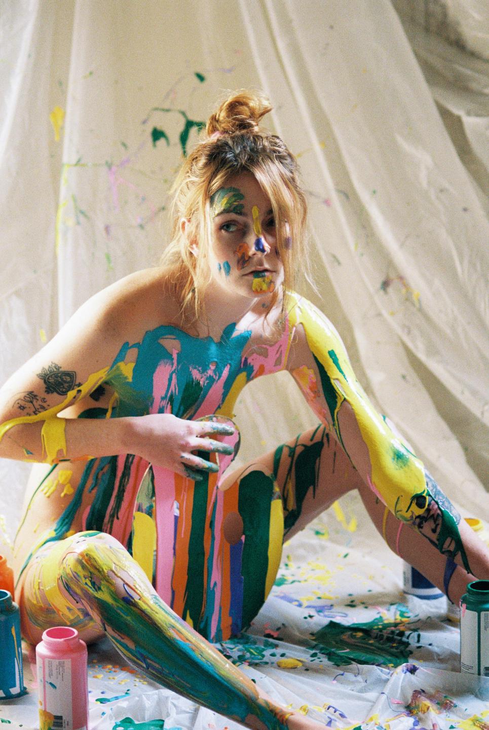 A woman sits covered in colorful paint with paint splatters on the white sheet behind her.