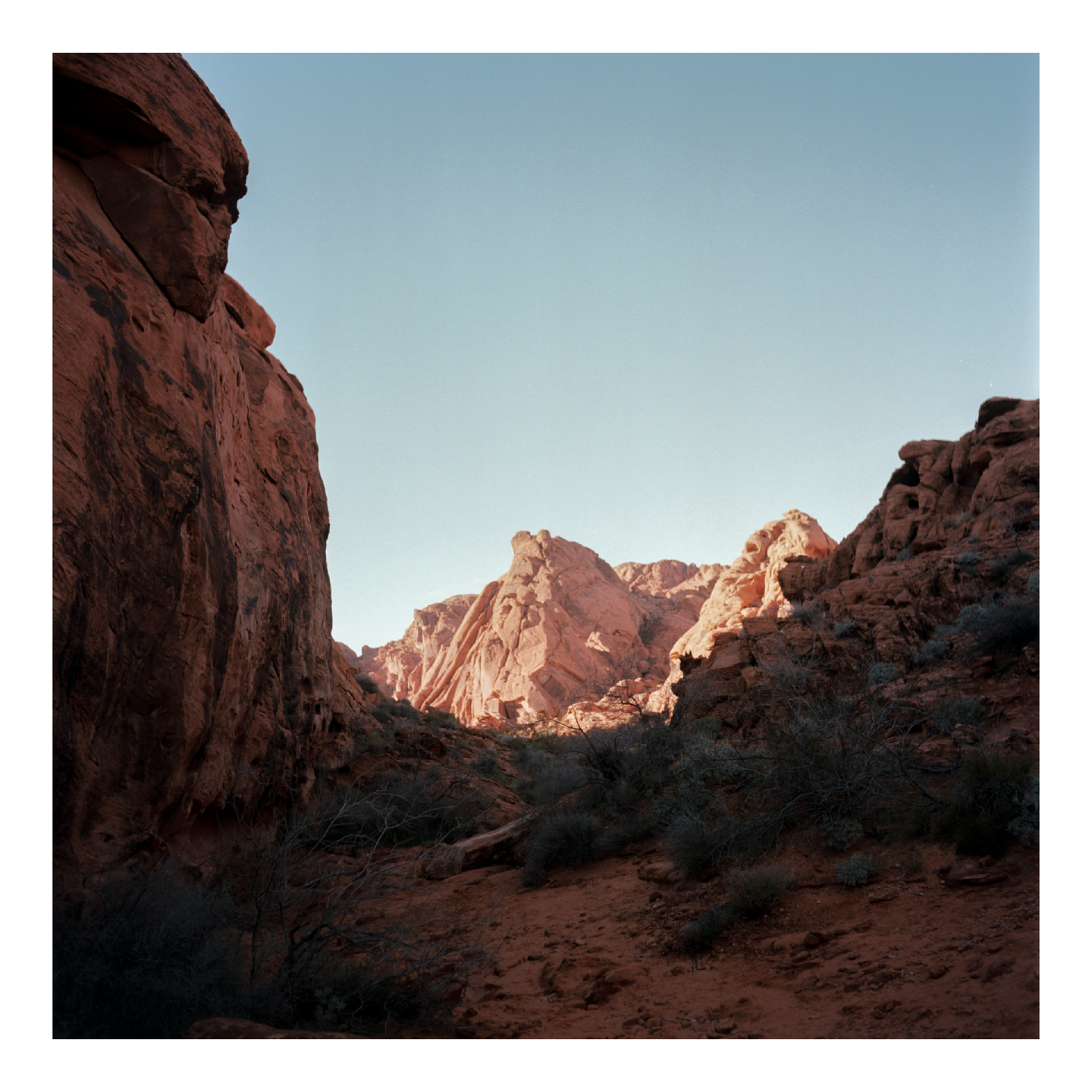 A desert landscape features red rock formations under a clear sky, with some areas in shadow and others illuminated by sunlight.