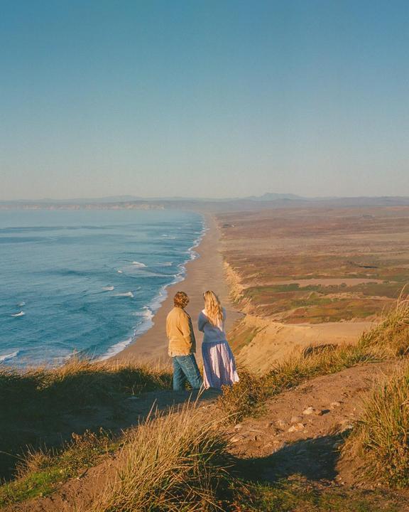A couple stands on a grassy cliff overlooking a long sandy beach and the ocean.