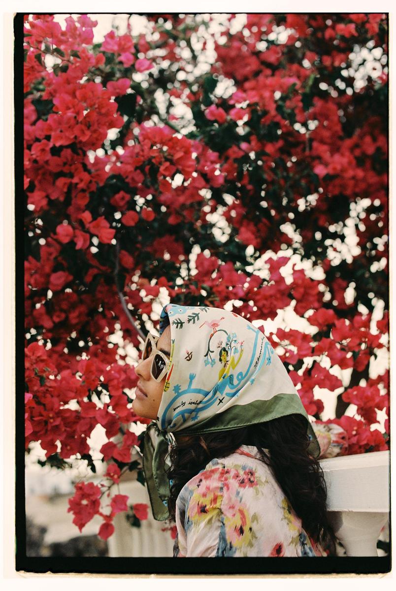 A woman wearing a headscarf and sunglasses looks up at a vibrant bougainvillea bush.