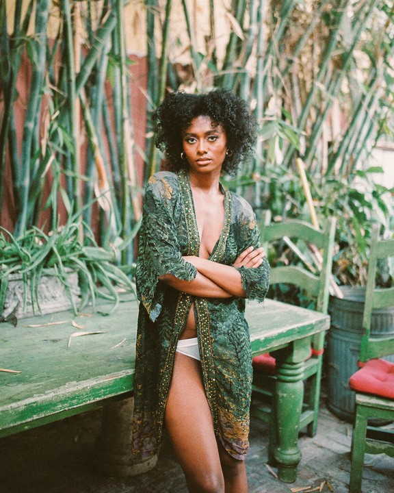A woman with curly hair stands with her arms crossed, wearing an open robe and underwear, next to a green wooden table with bamboo in the background.