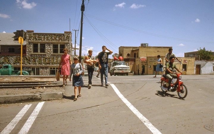 A group of people walk across a street with buildings and vehicles in the background.