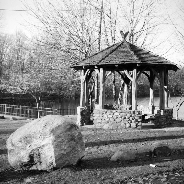 A gazebo with a stone base and wooden pillars stands by a body of water, surrounded by bare trees and large rocks in the foreground.