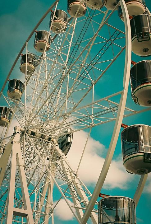 A white ferris wheel with multiple gondolas is set against a bright blue sky with white clouds.