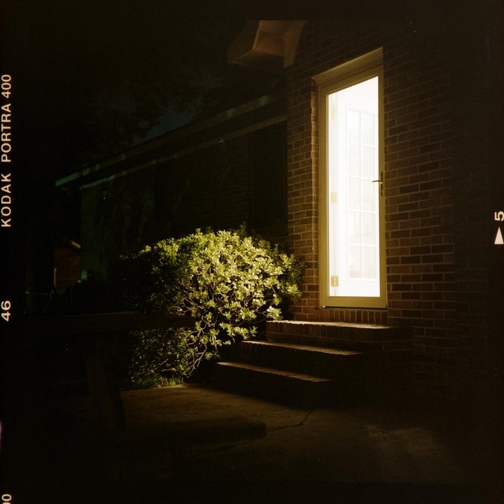 A brightly lit door and steps are next to a bush and a brick wall at night.