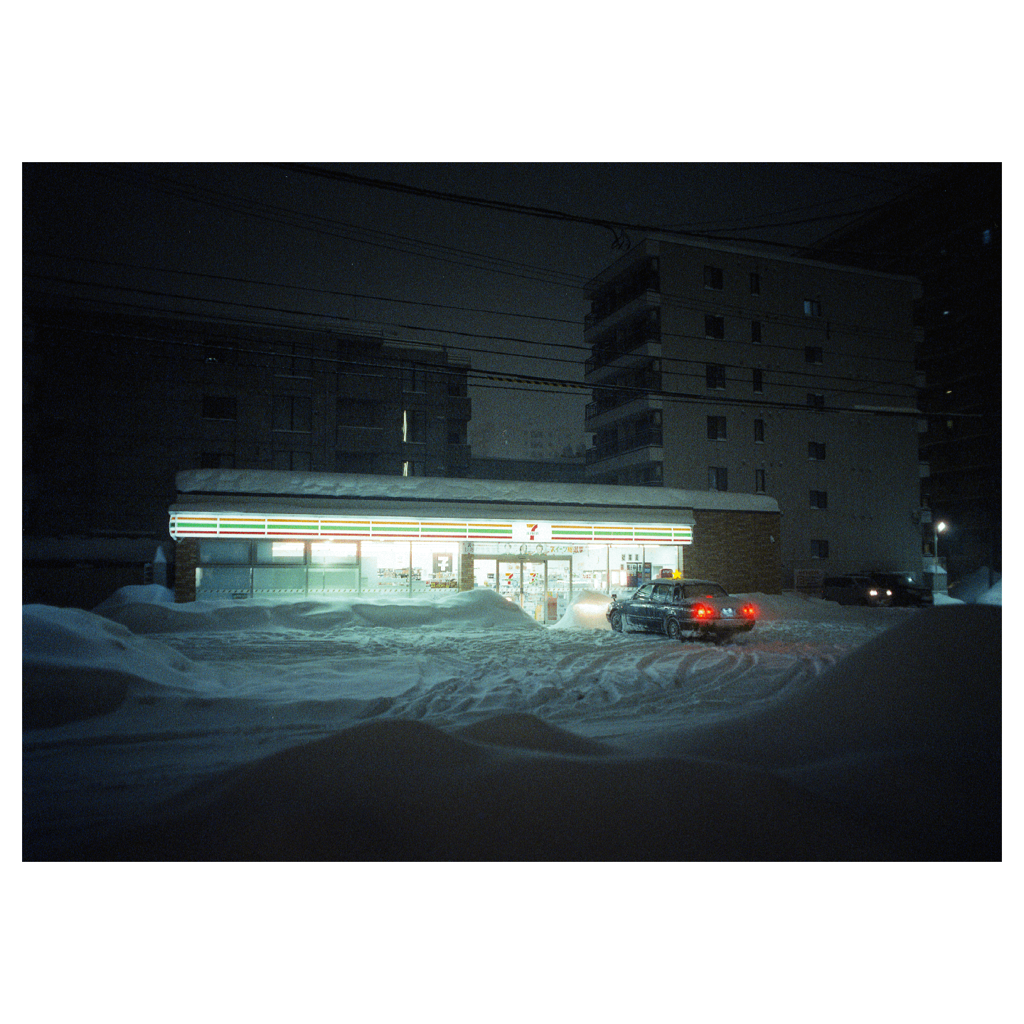 A 7-Eleven convenience store is brightly lit at night, surrounded by deep snow, with a car parked outside.