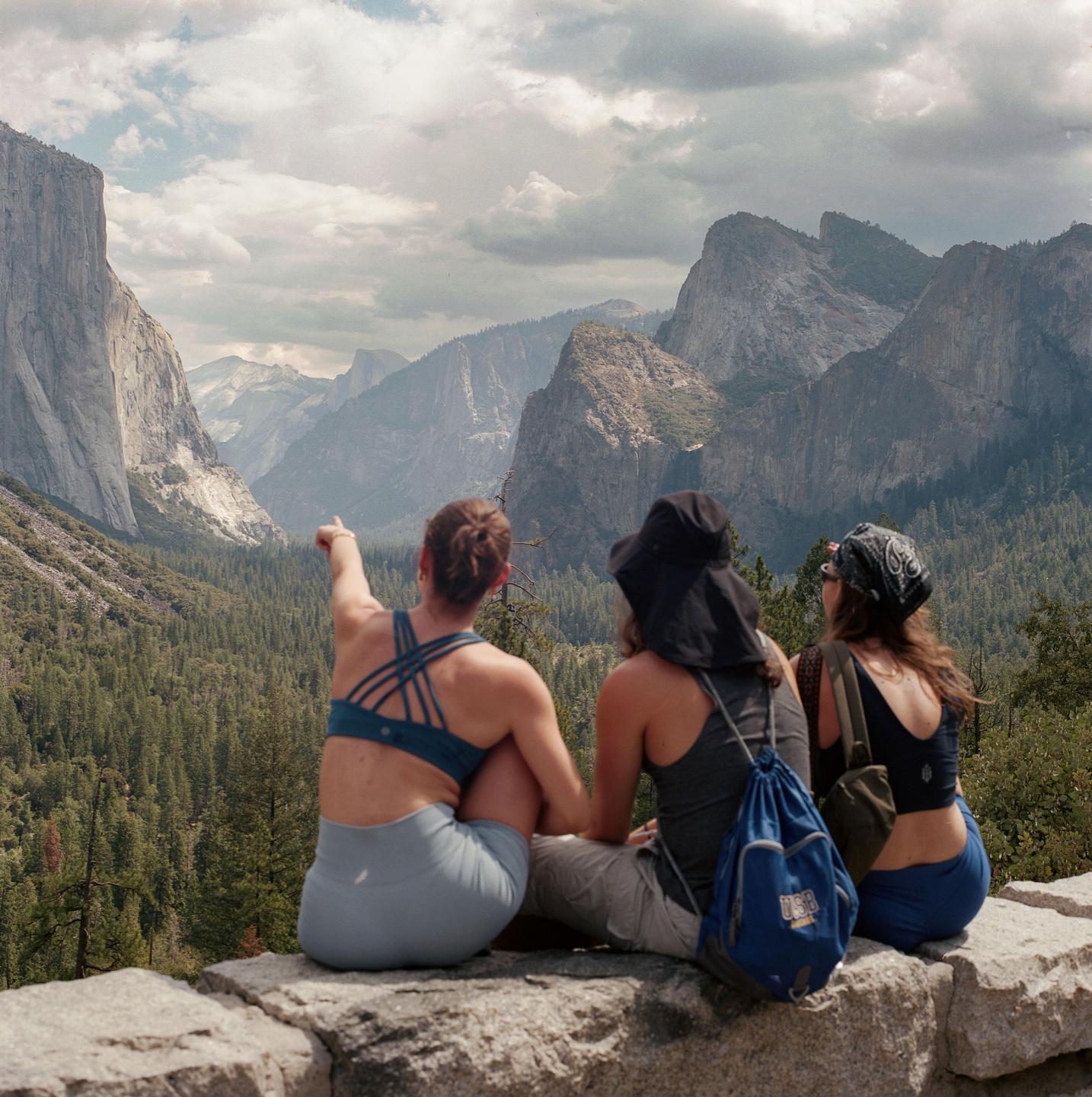 Three women sit on a stone wall overlooking a valley with mountains and a forest.