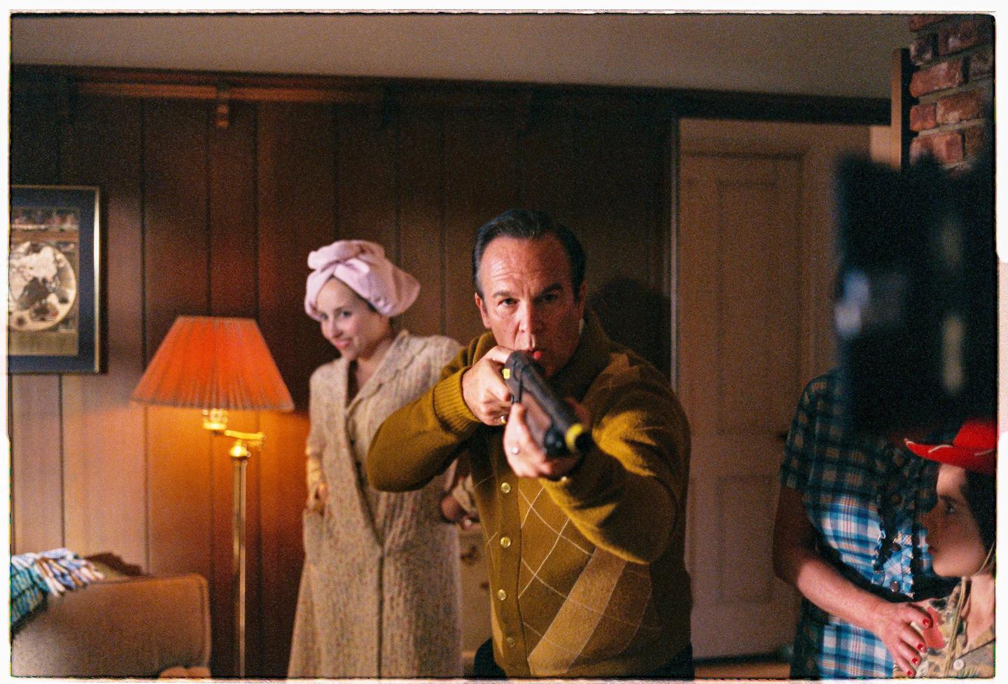 A man aims a shotgun at the viewer while a woman in a bathrobe and a child in a cowboy hat stand behind him.