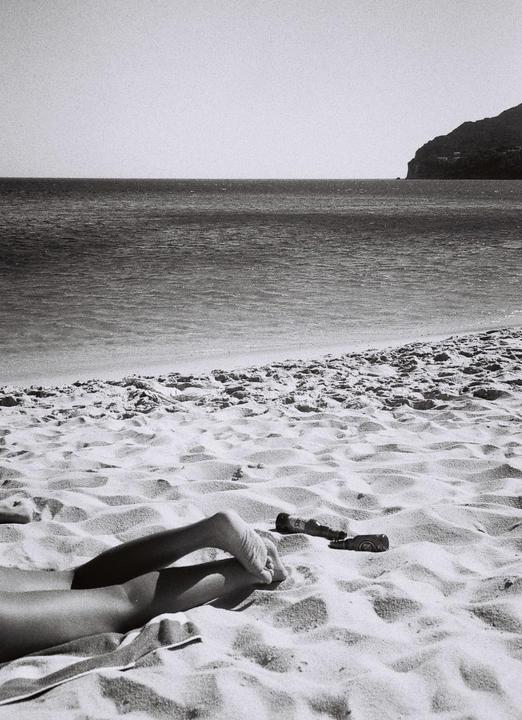 A person's legs and feet are visible on a sandy beach with the ocean and a distant mountain in the background.