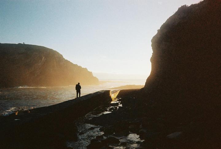A man stands on a rocky outcrop looking out at the ocean between two large cliffs at sunset.